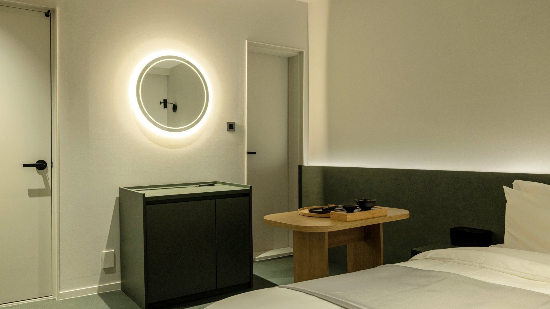 Una habitación de hotel moderna con una cama, una mesita de noche con espejo, una pequeña mesa de madera con tazas y un mueble de color oscuro.