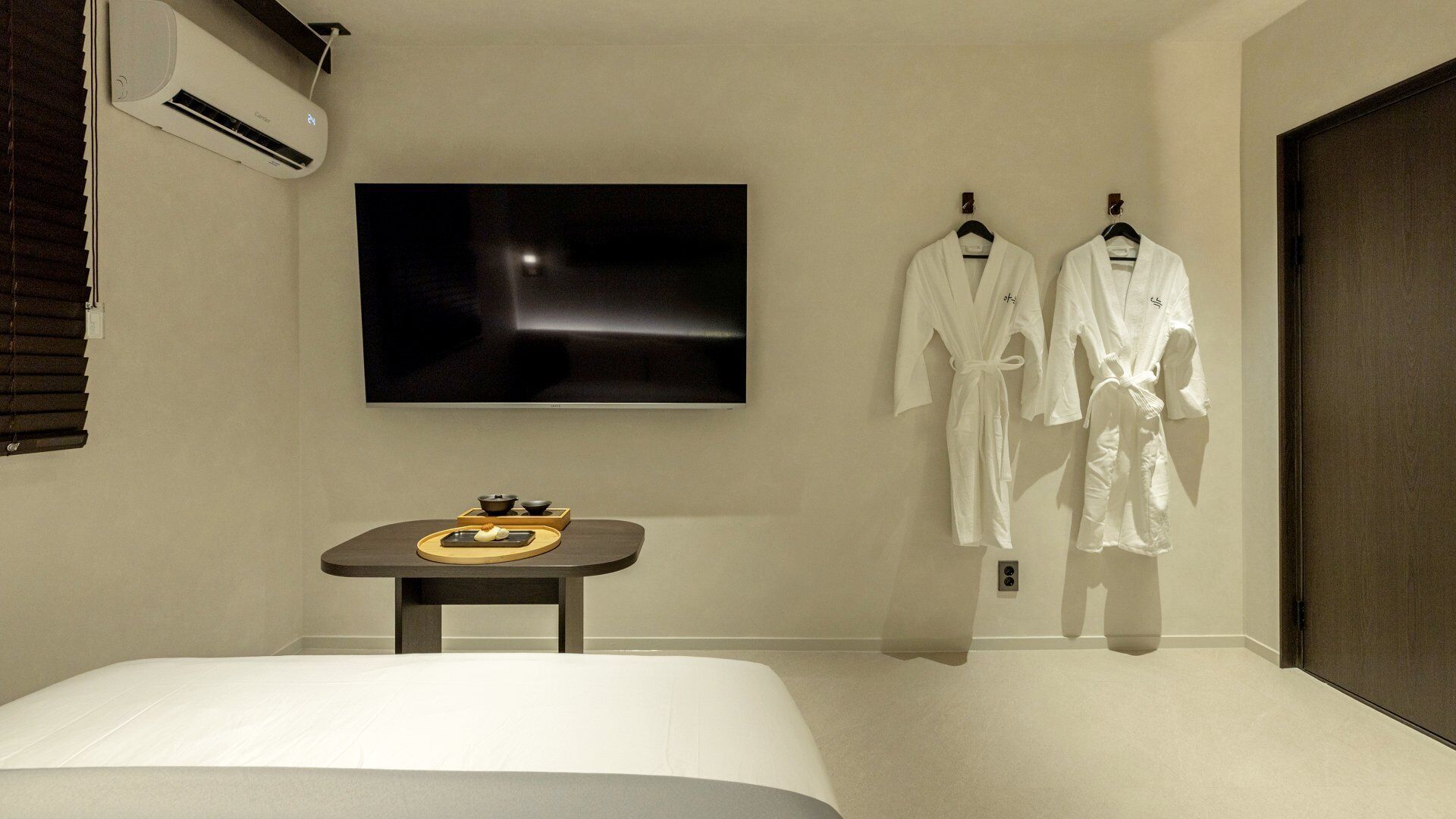 Habitación de hotel con una cama, un televisor de pantalla plana montado en la pared, una mesita con una bandeja y dos batas blancas colgadas en ganchos.