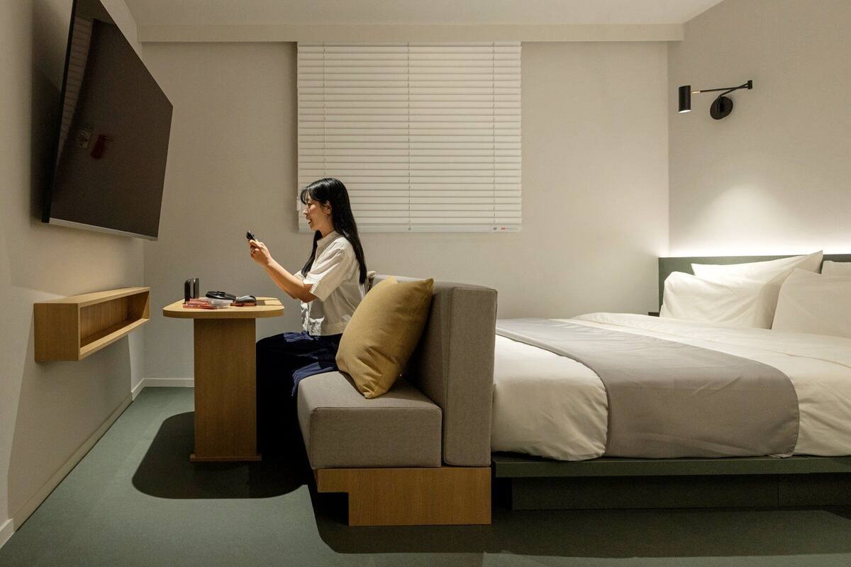 Une chambre d’hôtel avec un lit, un canapé, une petite table, une télévision et une fenêtre avec des stores.