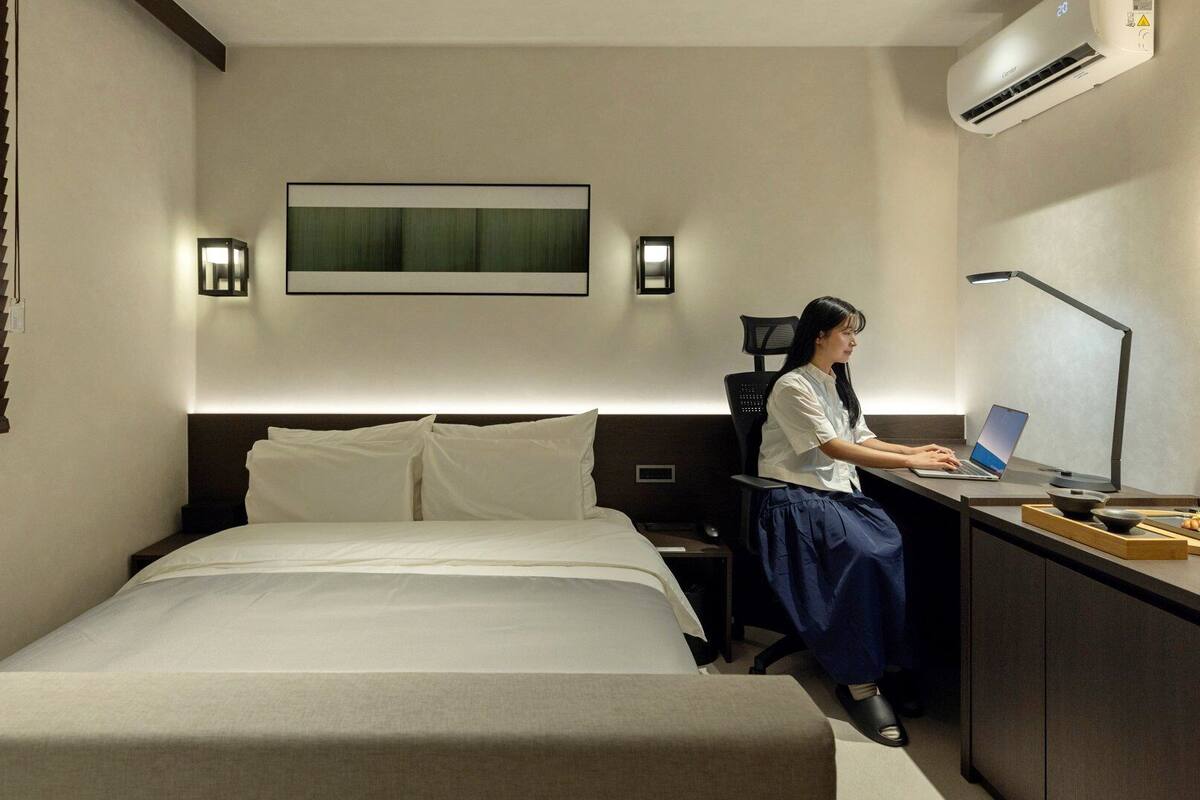 Une chambre d’hôtel équipée d’un lit, d’un bureau avec un ordinateur portable, d’une chaise et d’un climatiseur fixé au mur.
