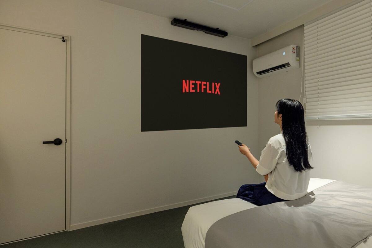 Une pièce comprenant un lit, une porte, un climatiseur fixé au mur et un grand écran affichant le mot « NETFLIX ».