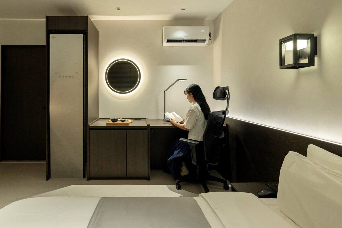 Une chambre d’hôtel comprenant un lit, un bureau avec une chaise, un climatiseur et une lampe fixée au mur.