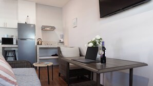 En kompakt tekøkken med køleskab, mikroovn og elkedel, et lille spisebord med en bærbar computer og en flaske, en sofa med en stribet pude, og et fjernsyn monteret på væggen.