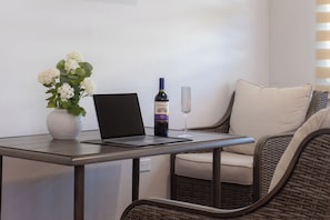 A laptop on a table with a bottle of wine and a glass, a vase with flowers, and a wicker chair.