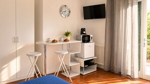 Habitación compacta con kitchenette, televisión de pantalla plana, reloj y puerta corrediza de vidrio al exterior.