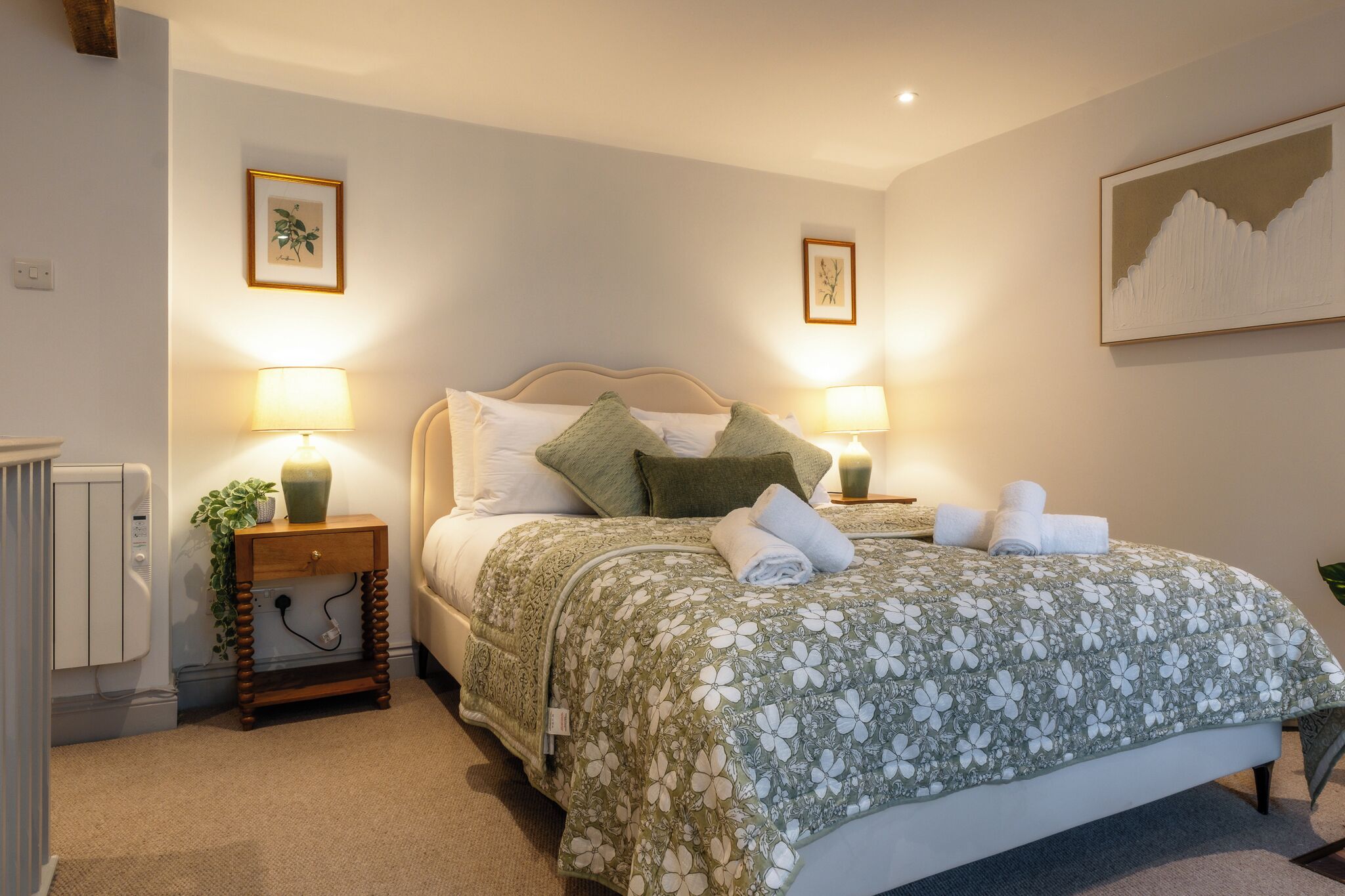 A neatly made bed with a floral bedspread, two bedside lamps, a small bedside table with a plant, and a framed picture on the wall.