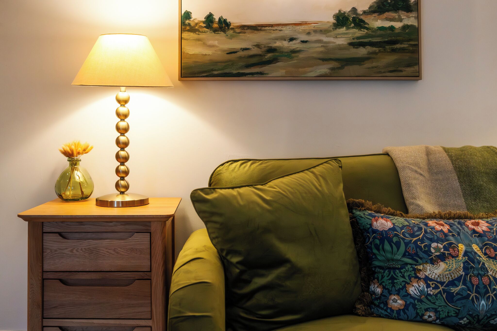 A green sofa with a floral pillow, a lamp with a vase of flowers, and a painting on the wall.