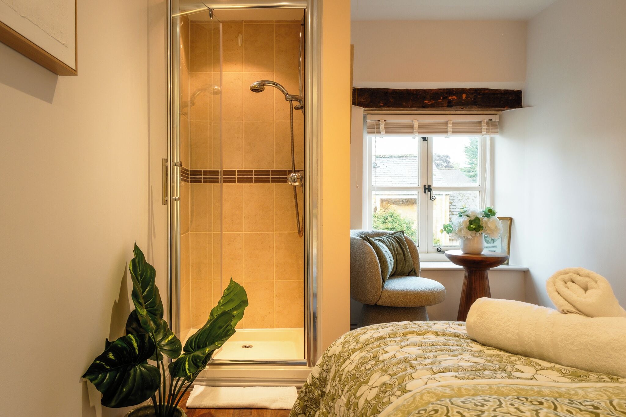 A bathroom with a glass-enclosed shower, a bed with a patterned bedspread, a window with a view, and a small table with a plant.