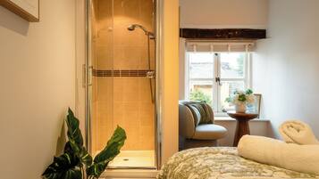A bathroom with a glass-enclosed shower, a bed with a patterned bedspread, a window with a view, and a small table with a plant.