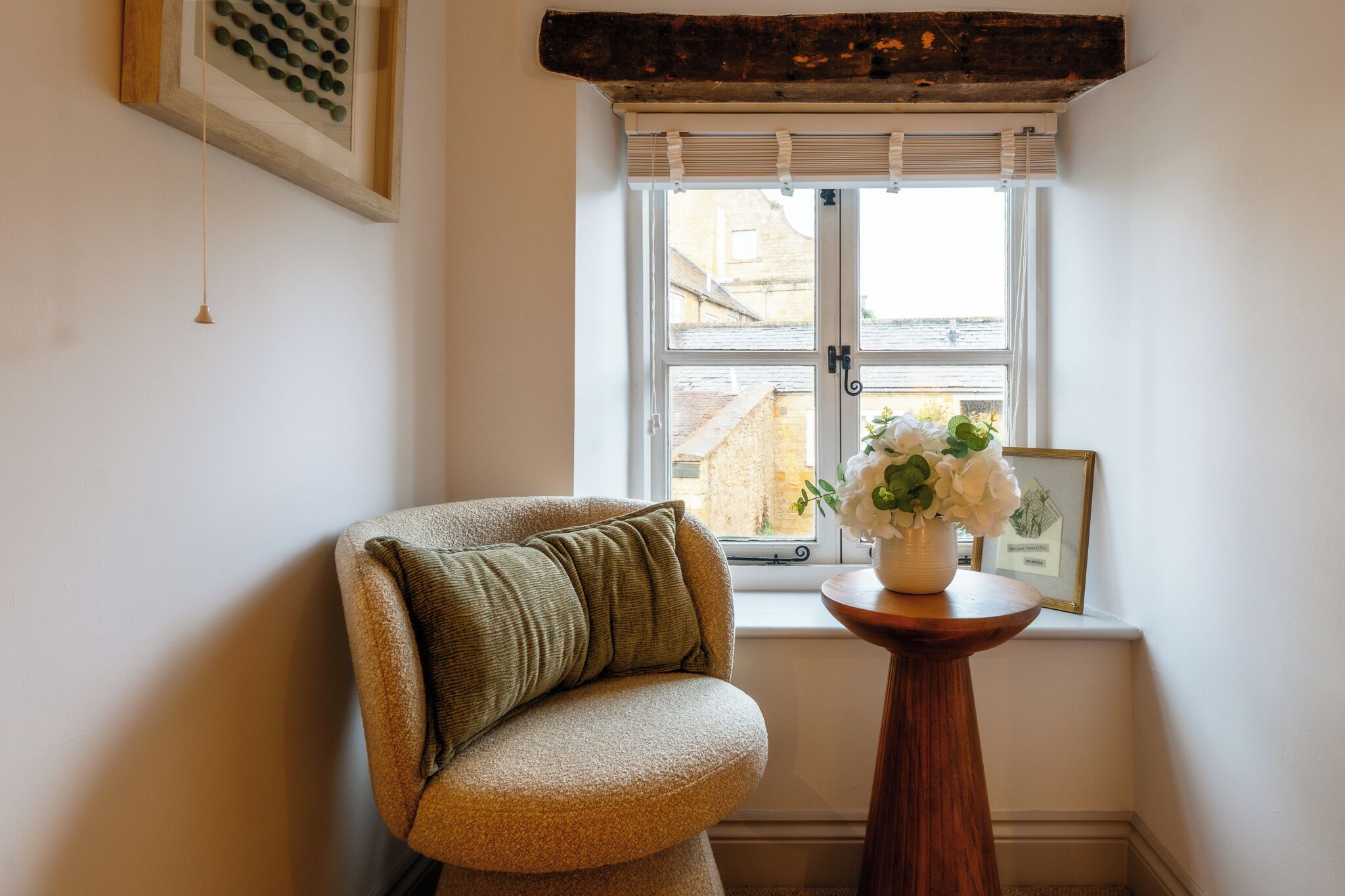 Ein Eckzimmer mit gepolstertem Sessel, einem kleinen Holztisch mit Blumenvase und einem Fenster mit Jalousien. An der Wand hängt ein gerahmtes Bild.