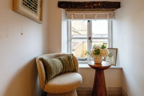 A corner room with a cushioned chair, a small wooden table with a vase of flowers, a window with blinds, and a framed picture on the wall. - Little Cottage in Moreton-in-Marsh (Moreton-in-Marsh)