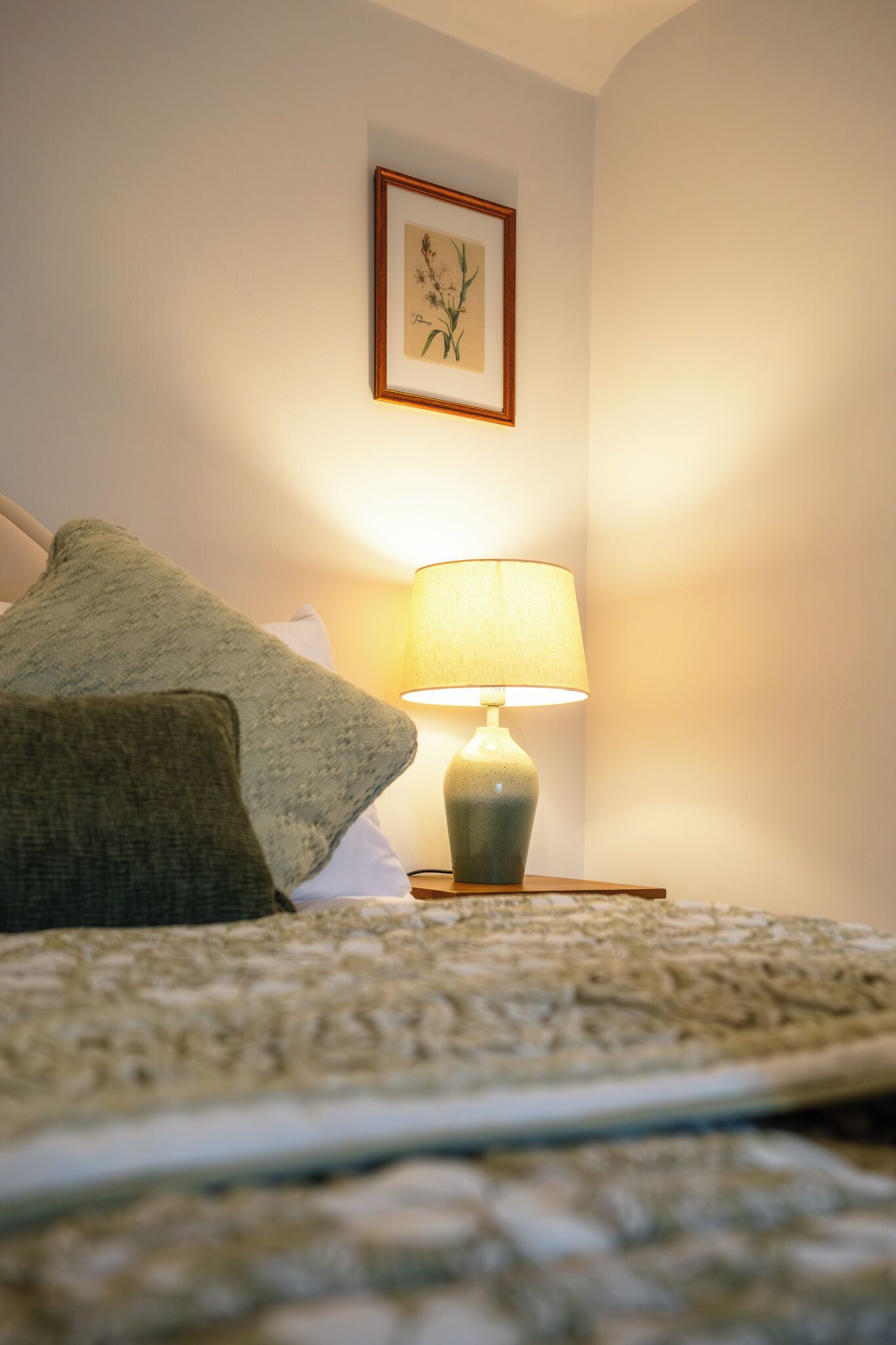 A bed with a green pillow, a bedside lamp, and a framed botanical print on the wall.
