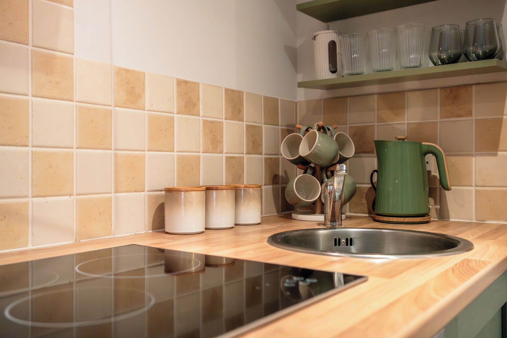A kitchen with a sink, a green kettle, and a shelf with cups and containers.