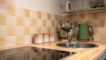 A kitchen with a sink, a green kettle, and a shelf with cups and containers.