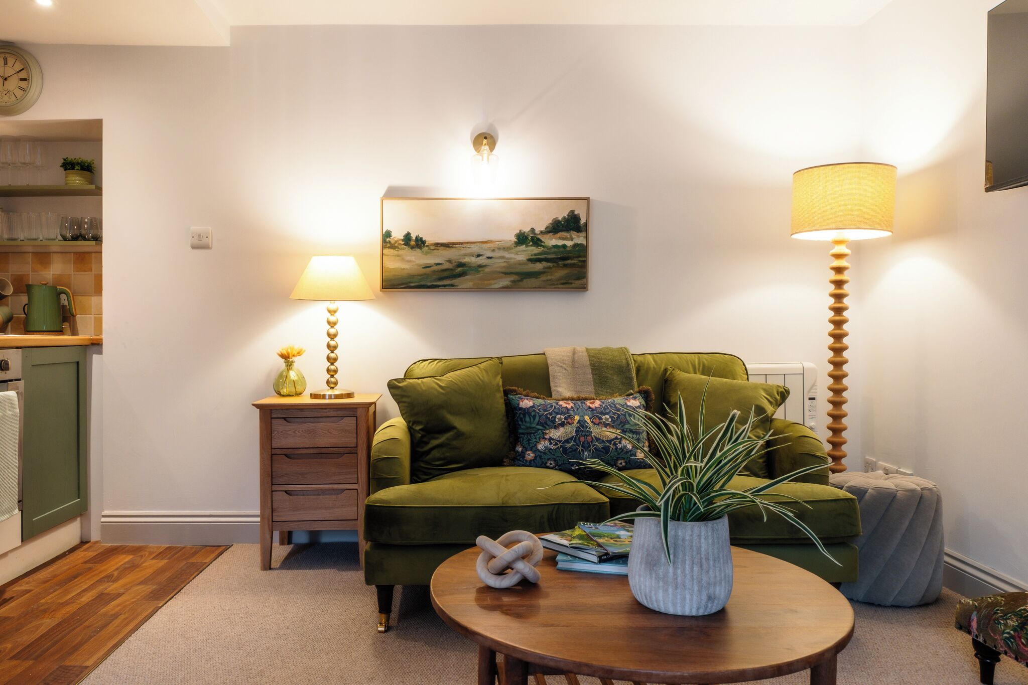 Ein gemütliches Wohnzimmer mit einem grünen Sofa, einem Holztisch, einem Bild an der Wand und einer Lampe.
