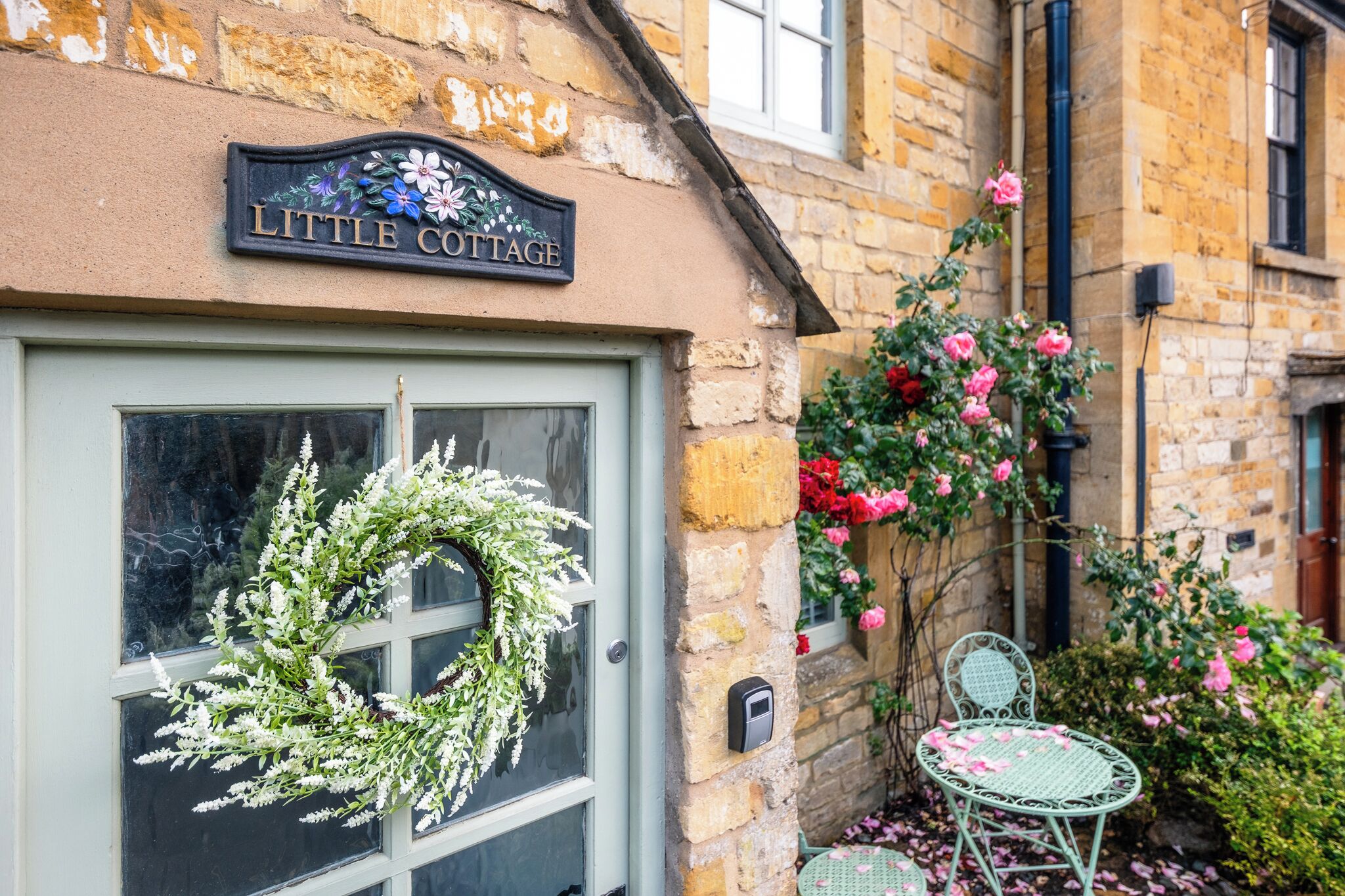 A stone building with a sign reading 'Little Cottage,' a door with a wreath, and a small outdoor seating area with chairs.