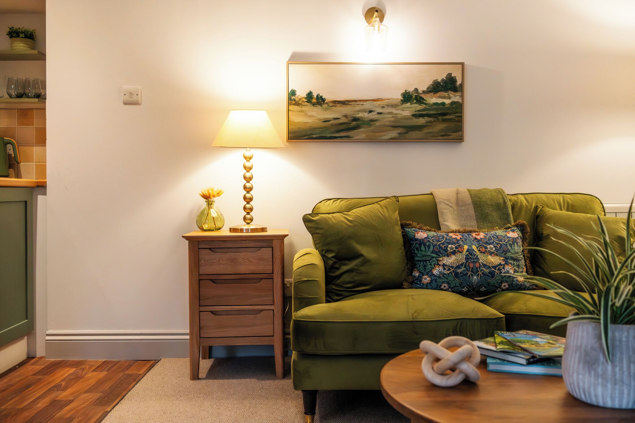 Ein grünes Sofa, ein Nachttisch aus Holz mit einer Lampe, ein Bild an der Wand und ein Blumentopf.