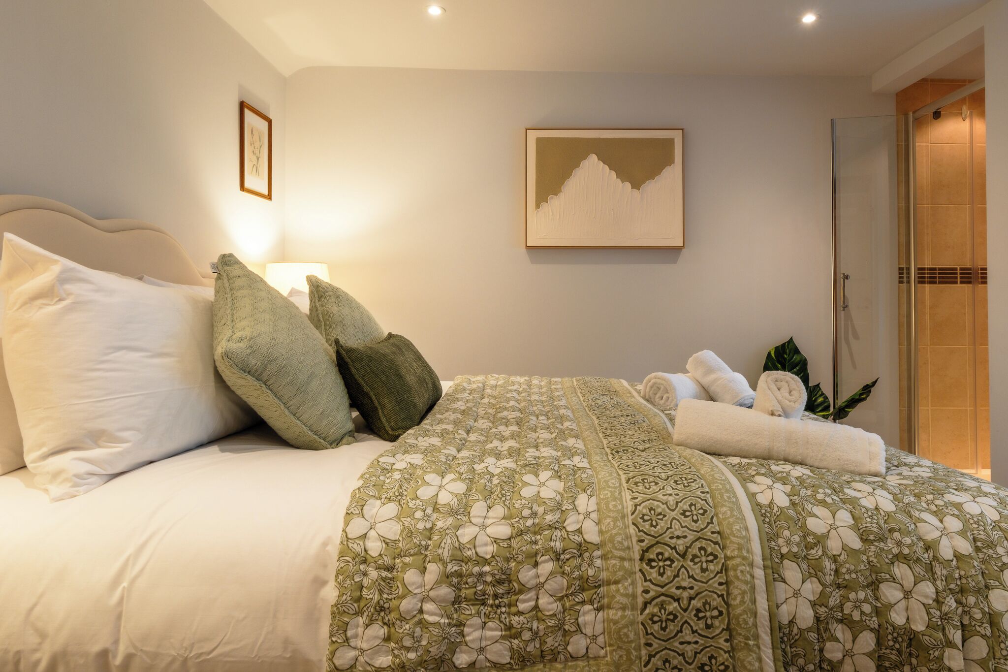 Un lit bien fait, recouvert d’une couvre-lit à motifs floraux, un tableau accroché au mur et une porte donnant sur une salle de bain.
