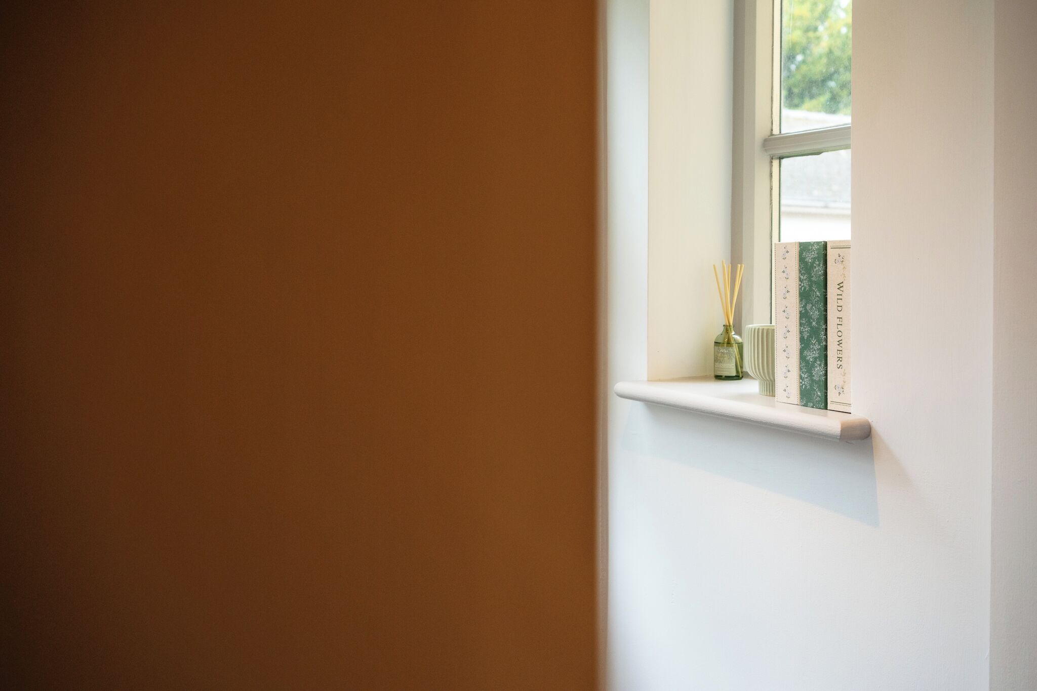 Ein Fenster mit Ablage, auf der ein Buch mit dem Titel 'Wild Flowers' und eine kleine Vase mit einer Kerze stehen.