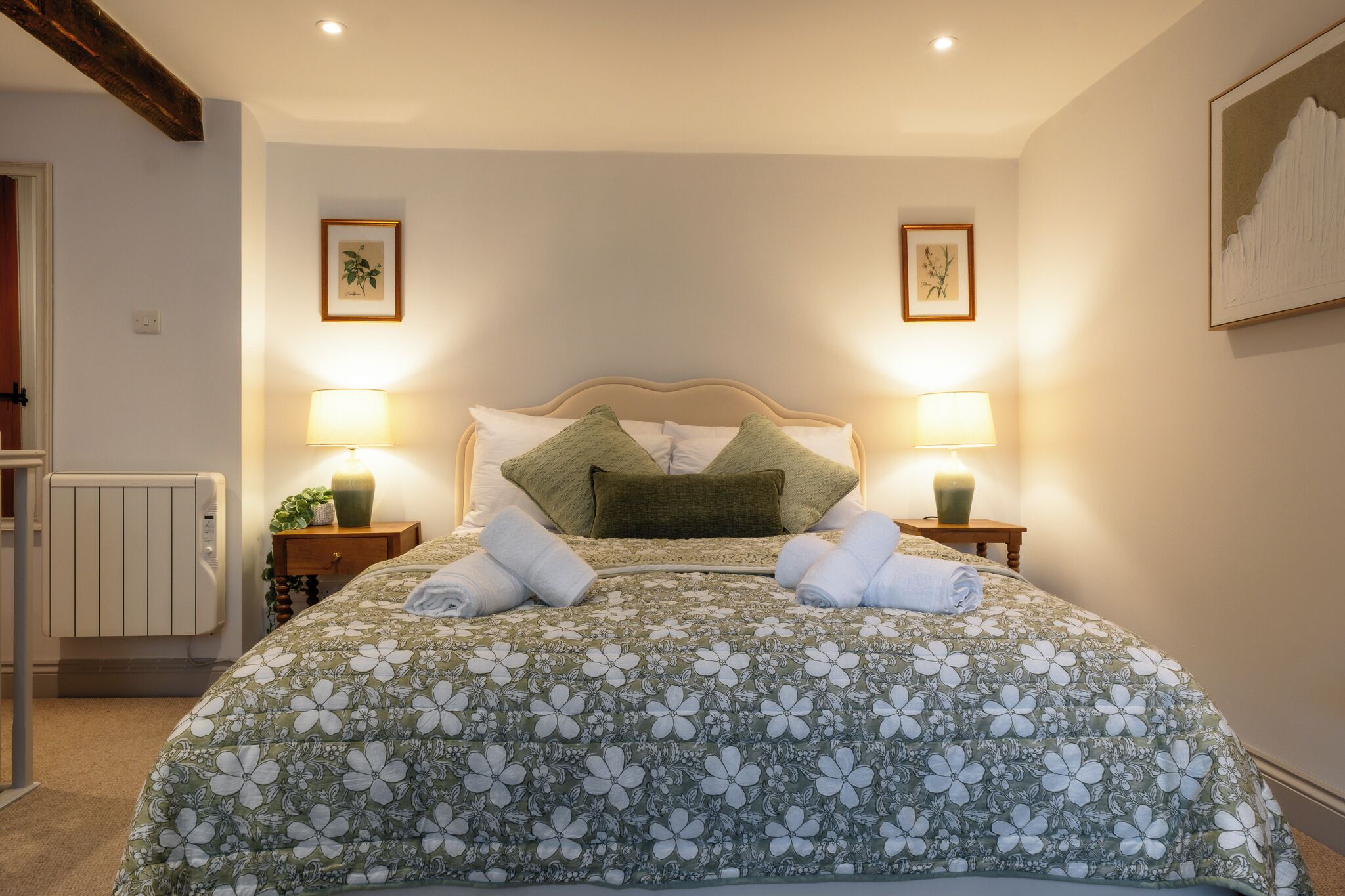 A bedroom with a floral bedspread, two bedside lamps, a radiator, and framed artwork on the walls.