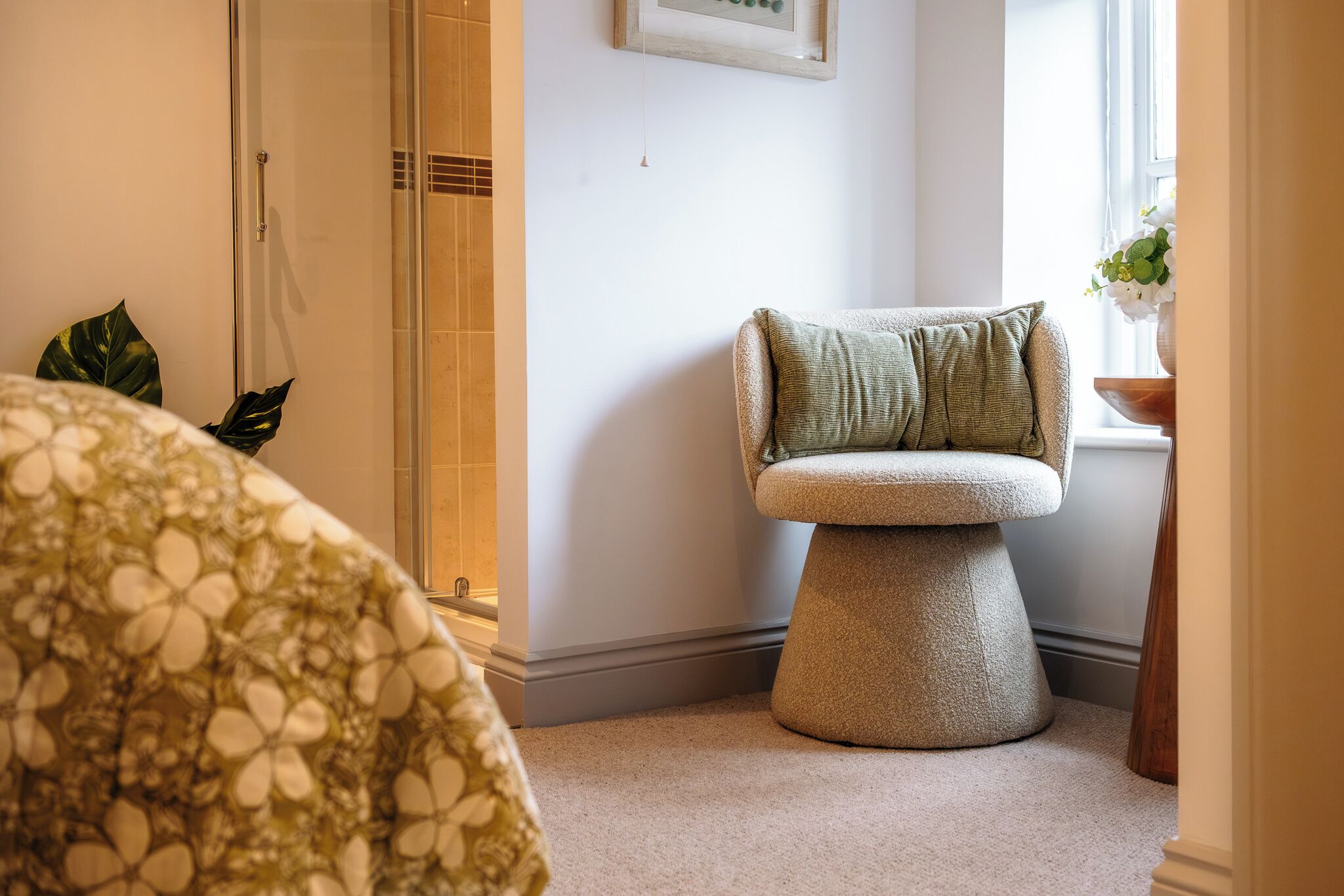 A corner of a room with a glass shower, a round stool, a bed with a floral bedspread, and a window with a plant.