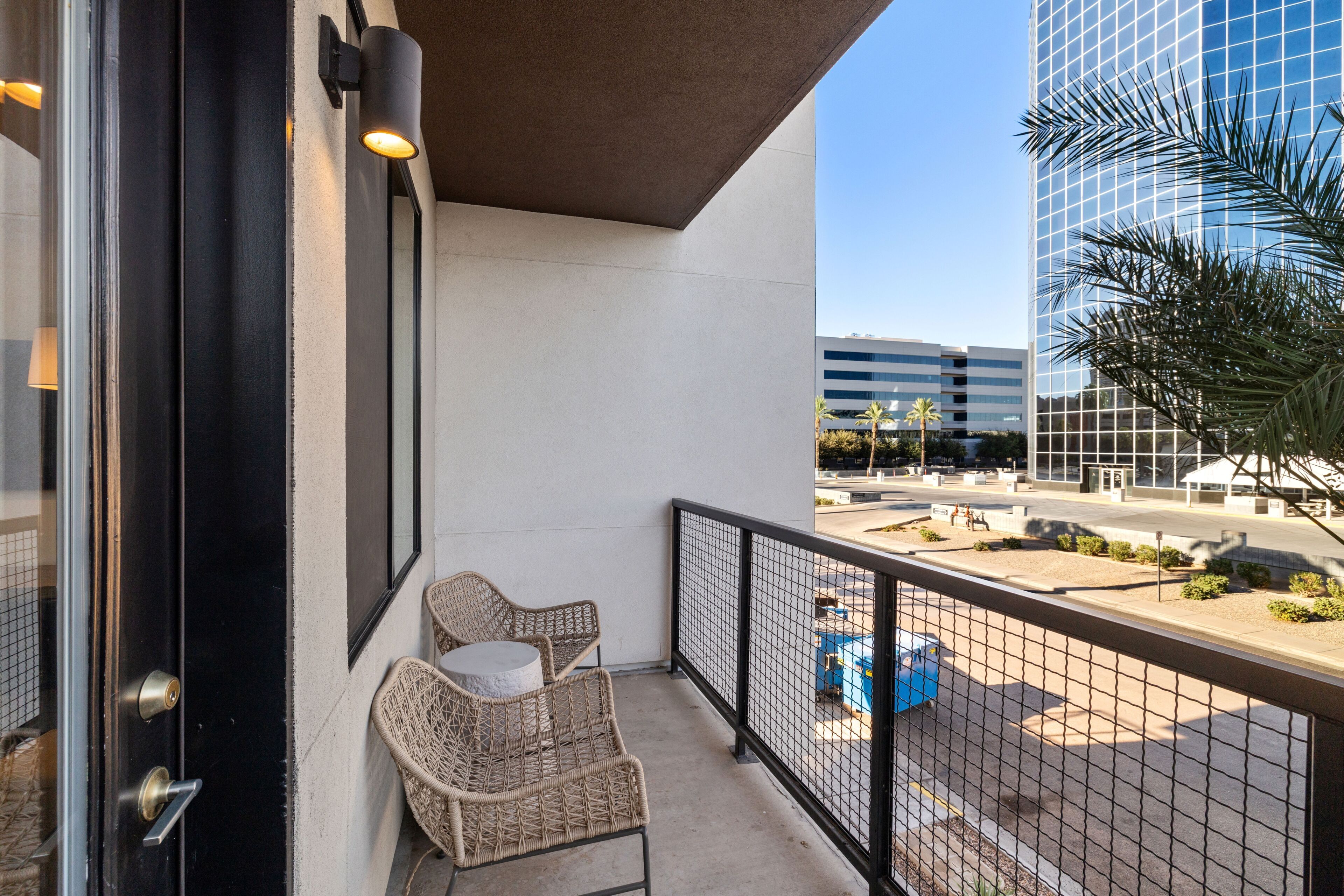 A balcony with wicker chairs, a glass door, and a view of a modern building and palm tree.