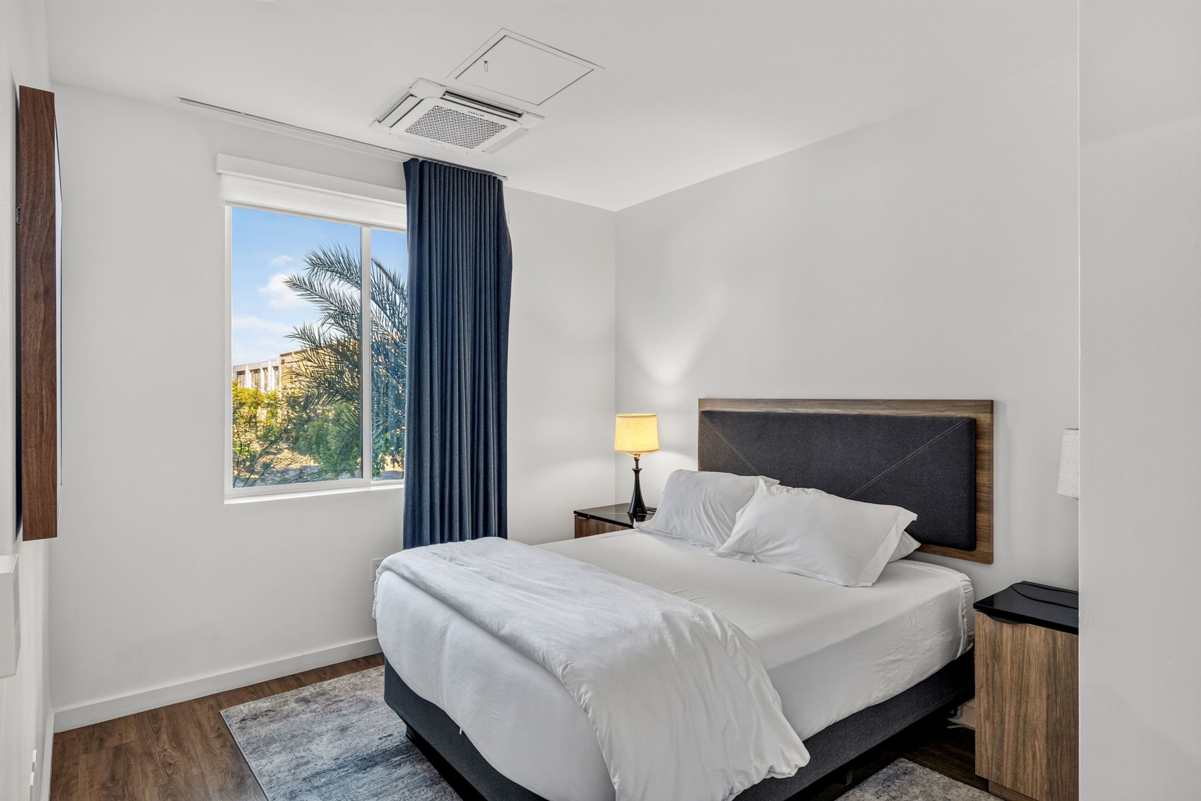 Una habitación de hotel moderna con una cama grande, una mesita de noche, una lámpara, una ventana con cortinas y vistas a palmeras.