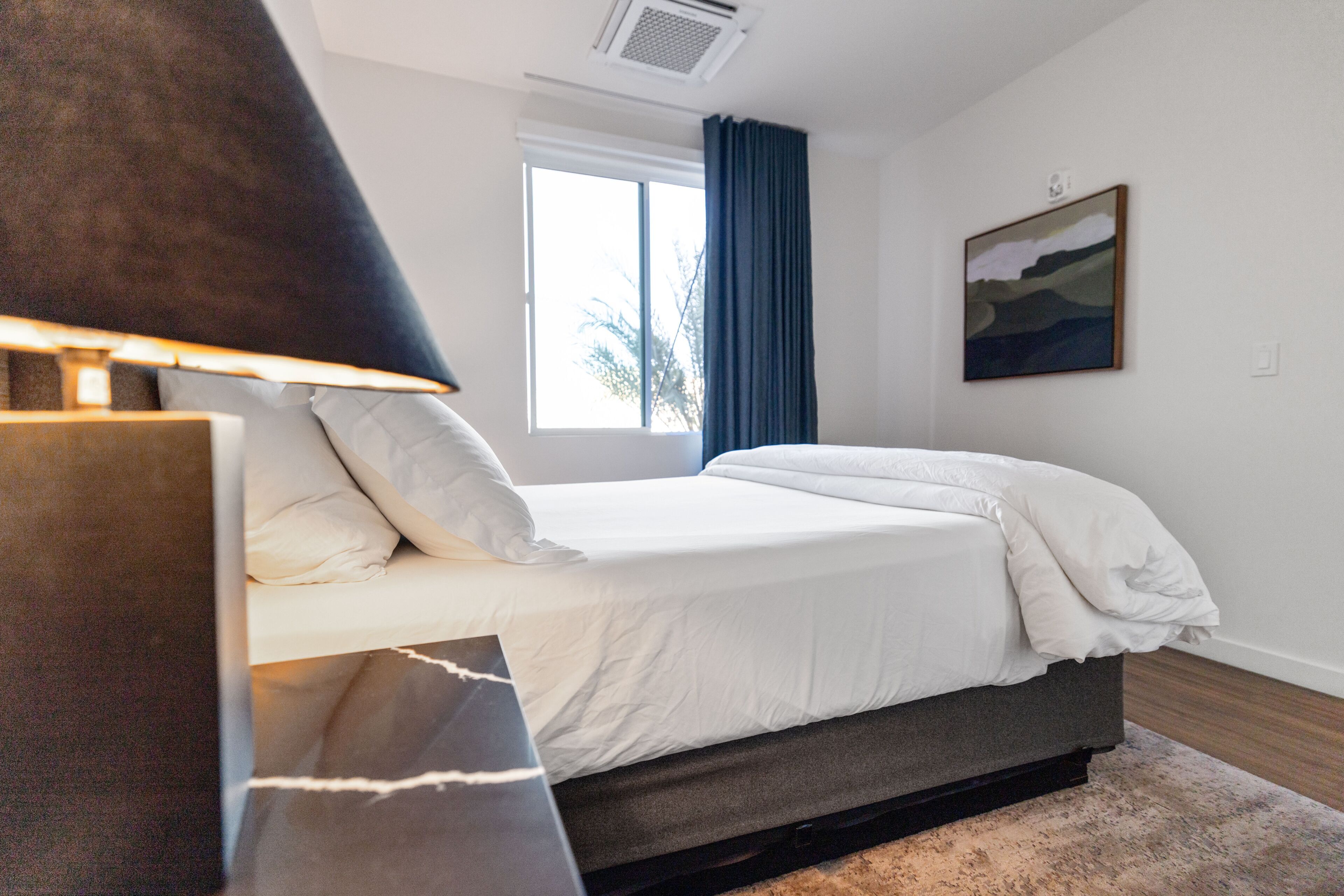 A bedroom with a bed, a bedside table, a lamp, a window with curtains, and a painting on the wall.