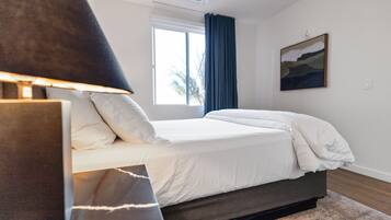 A bedroom with a bed, a bedside table, a lamp, a window with curtains, and a painting on the wall.