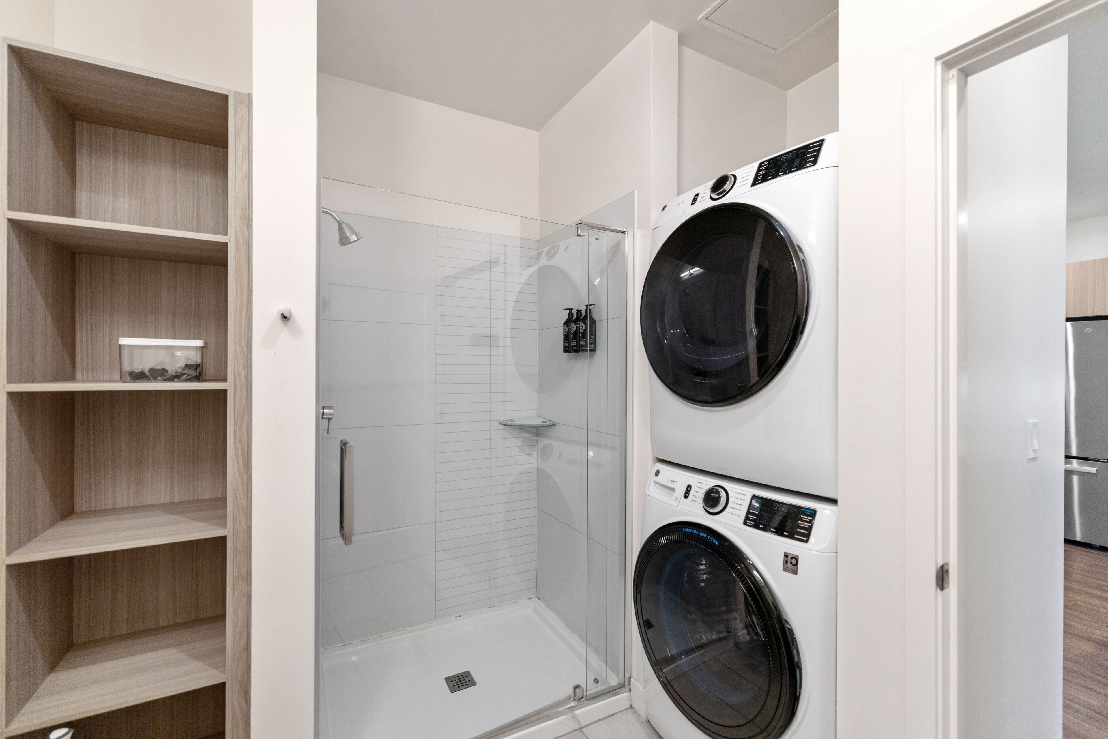 A modern laundry room with two front-loading washers, a glass-enclosed shower, and wooden shelving.