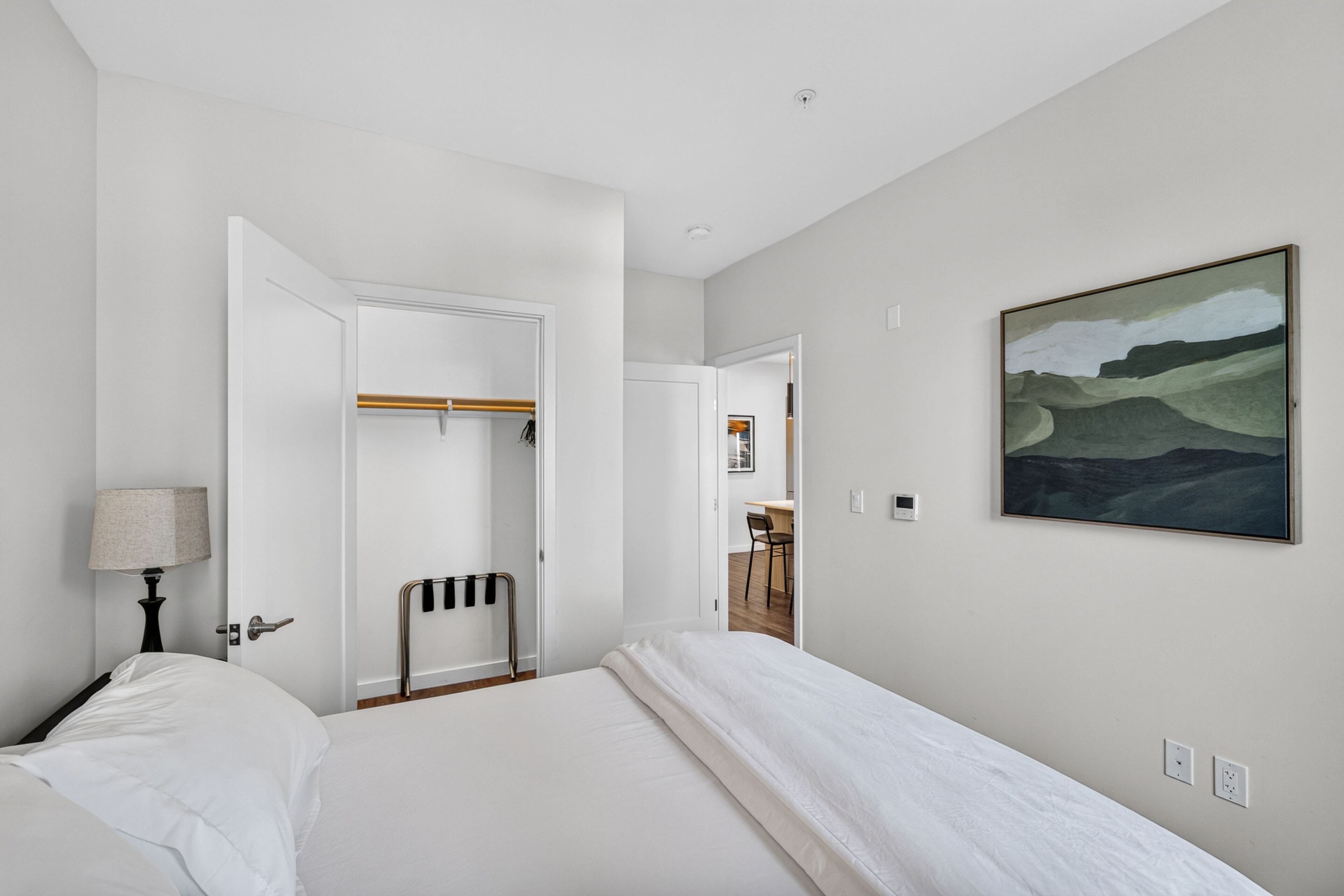 A bedroom with a bed, a bedside table with a lamp, a coat rack, and a painting on the wall.