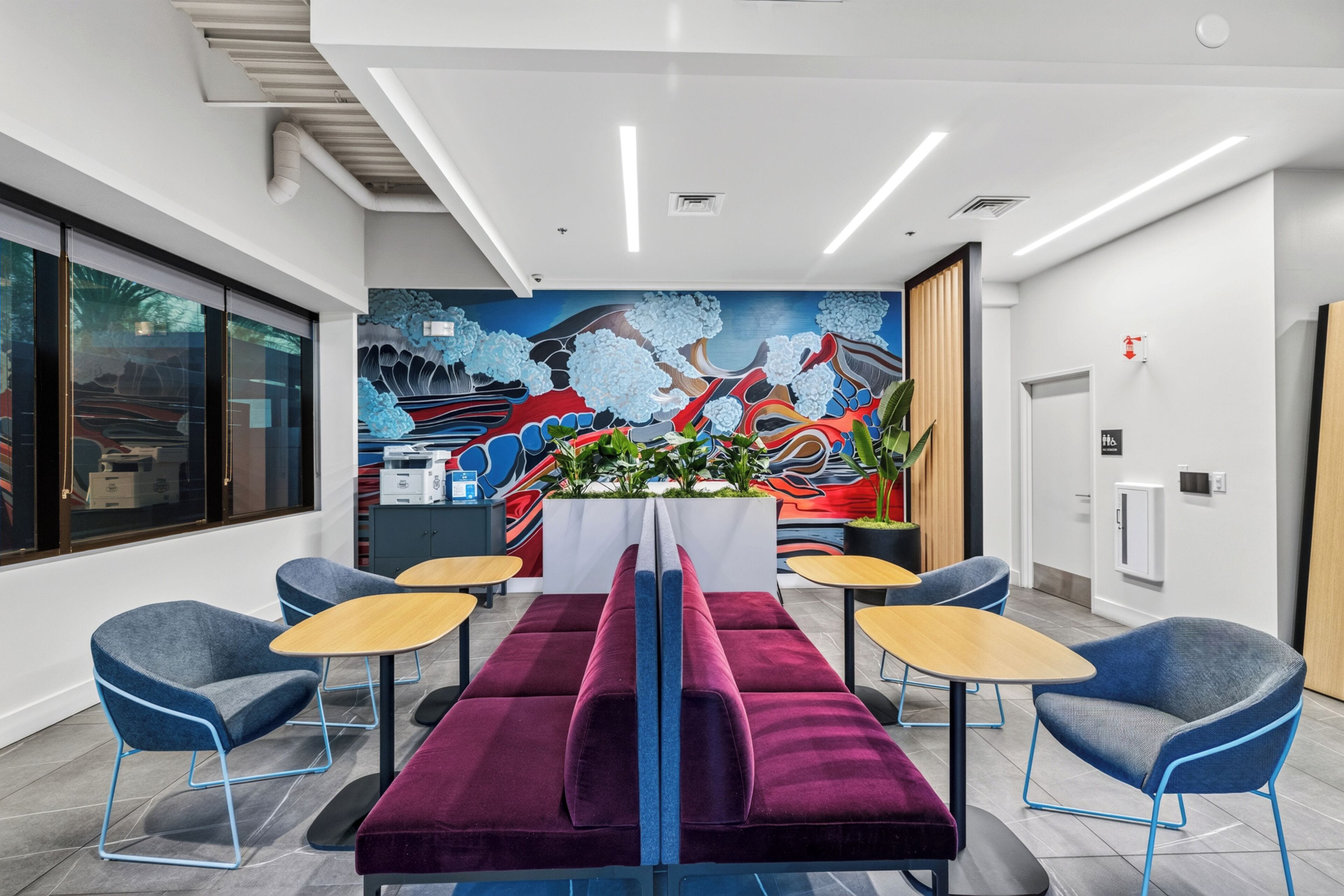 A modern hotel lobby with a long table, blue chairs, a colorful mural, and a reception desk.