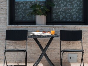 A table with a book, a glass of orange juice, and a plate with food, set between two chairs, against a brick wall with a window above. - Apartment Teide by Interhome (Pula)