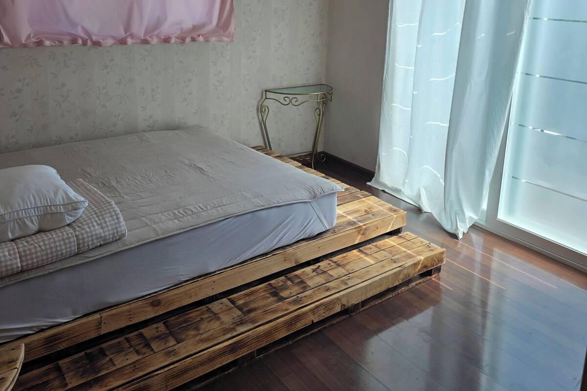 A bedroom with a bed on a wooden platform, a small metal table, a pink curtain, and a window with a white curtain.