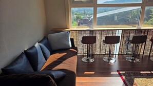 A living room with a sofa, three bar stools, a window with a view of a bridge and greenery, and a patterned rug. - MurMum 2 (Chilgok)