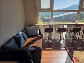 A living room with a sofa, three bar stools, a window with a view of a bridge and greenery, and a patterned rug.