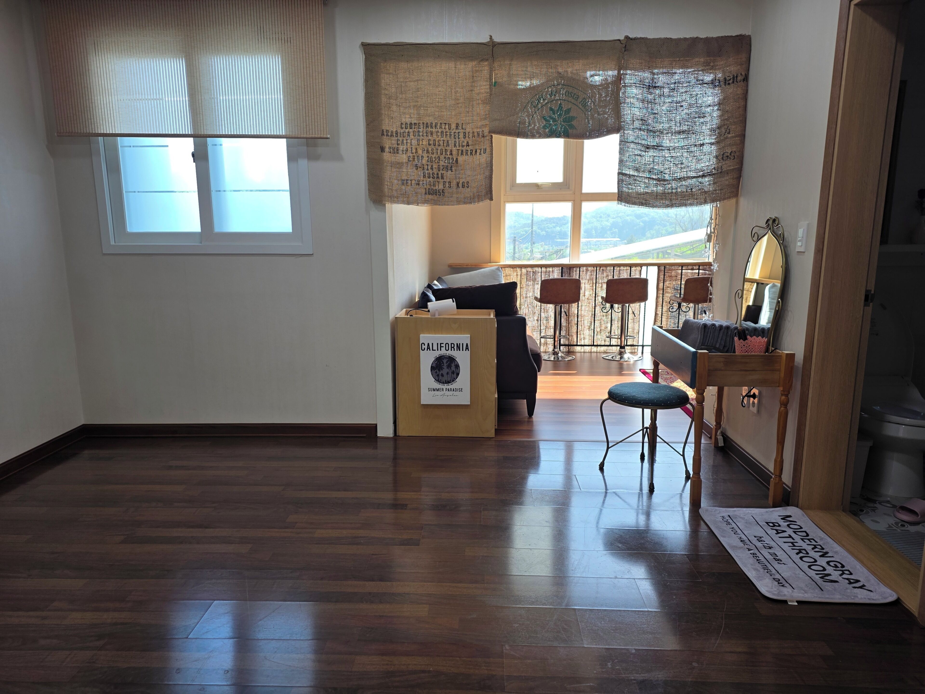 Una habitación con piso de madera, un sofá, una mesita, un taburete de bar, una ventana con persianas y vistas a un balcón.