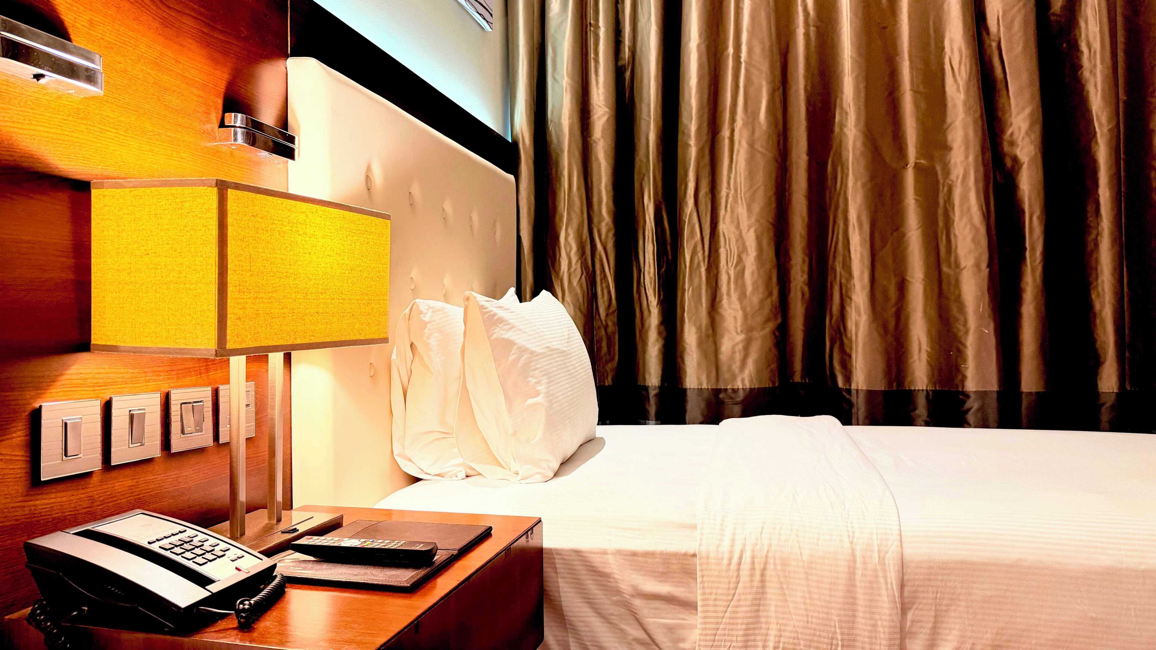 A hotel room with a bed, a desk with a telephone and a laptop, a lamp, and a window with curtains.