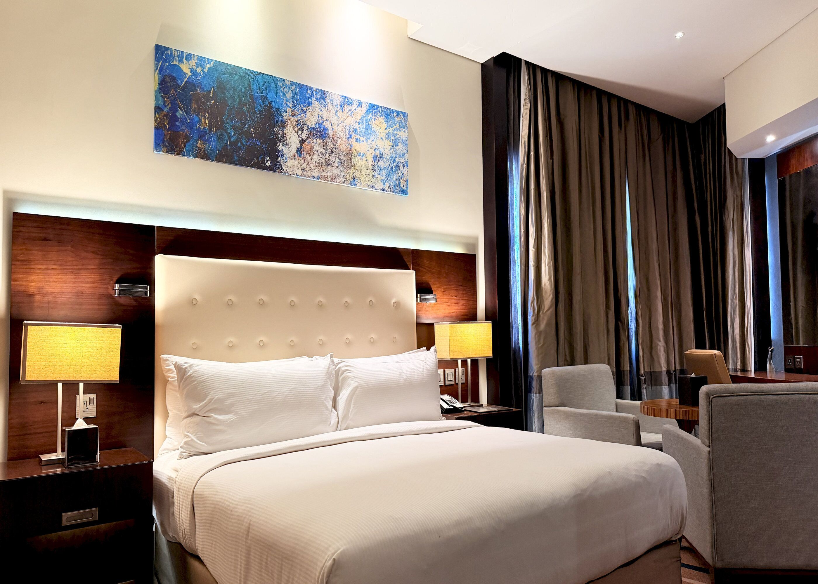 Habitación de hotel con una cama grande, dos mesitas de noche con lámparas, una silla, un escritorio y un cuadro abstracto de gran tamaño en la pared.