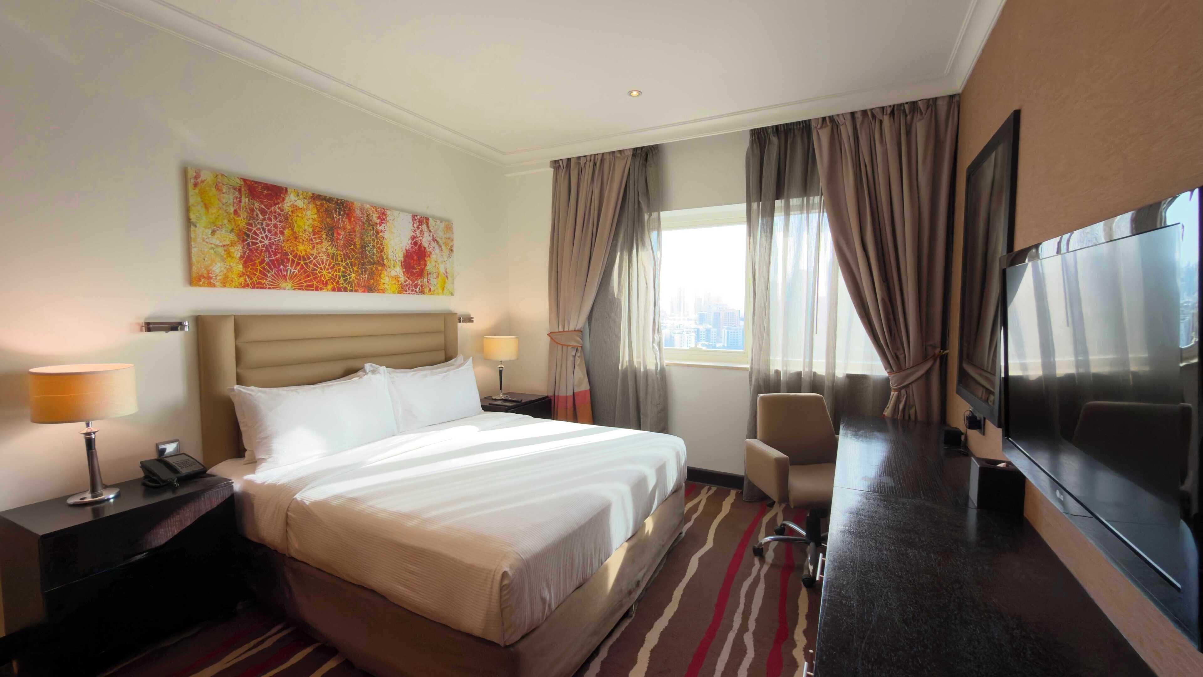 Habitación de hotel con cama, lámparas de noche, televisión, silla y un amplio ventanal con cortinas.