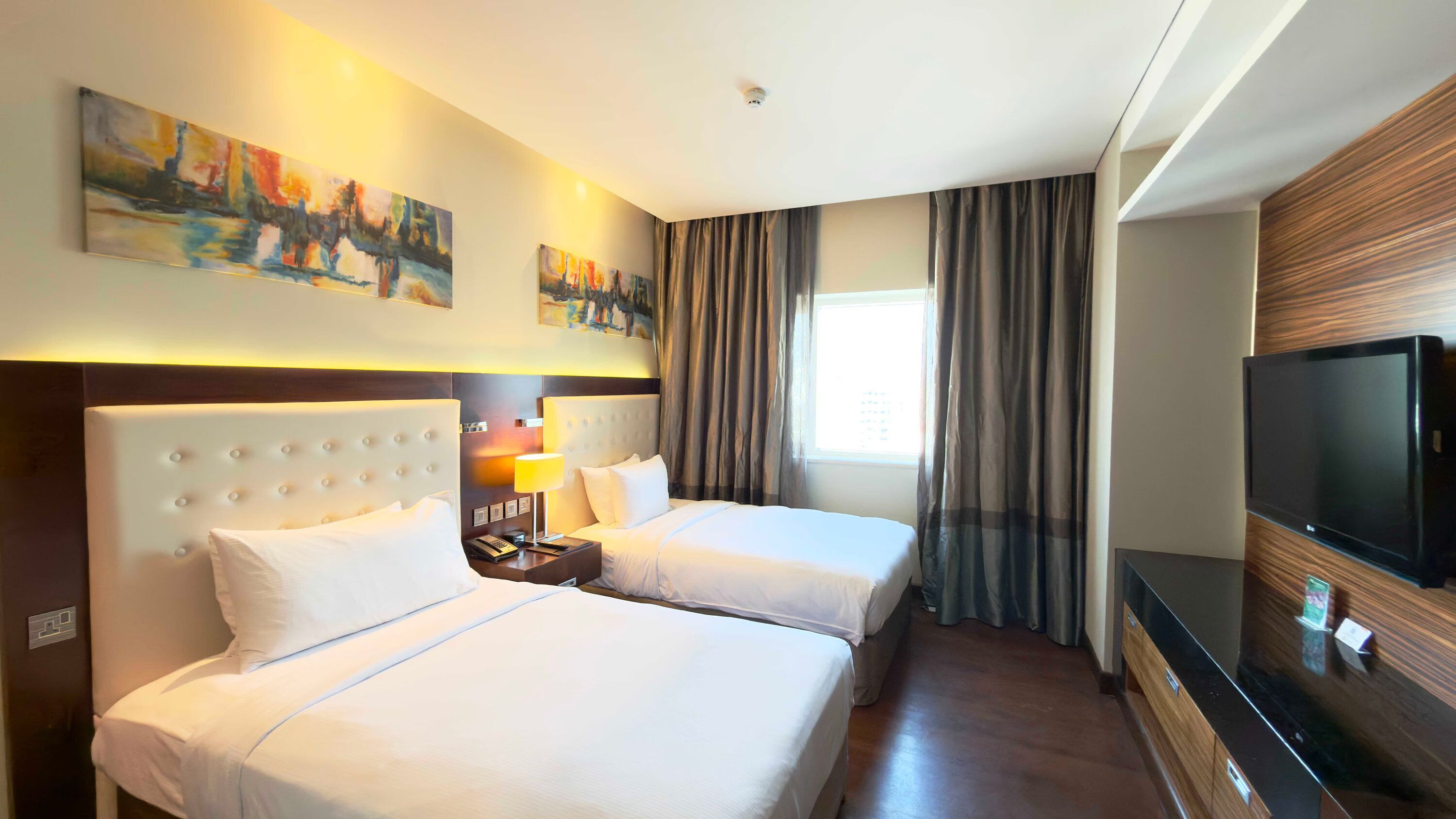 Ein Hotelzimmer mit zwei Betten, einem Fernseher, Kunstwerken an der Wand und einem Fenster mit Vorhängen.