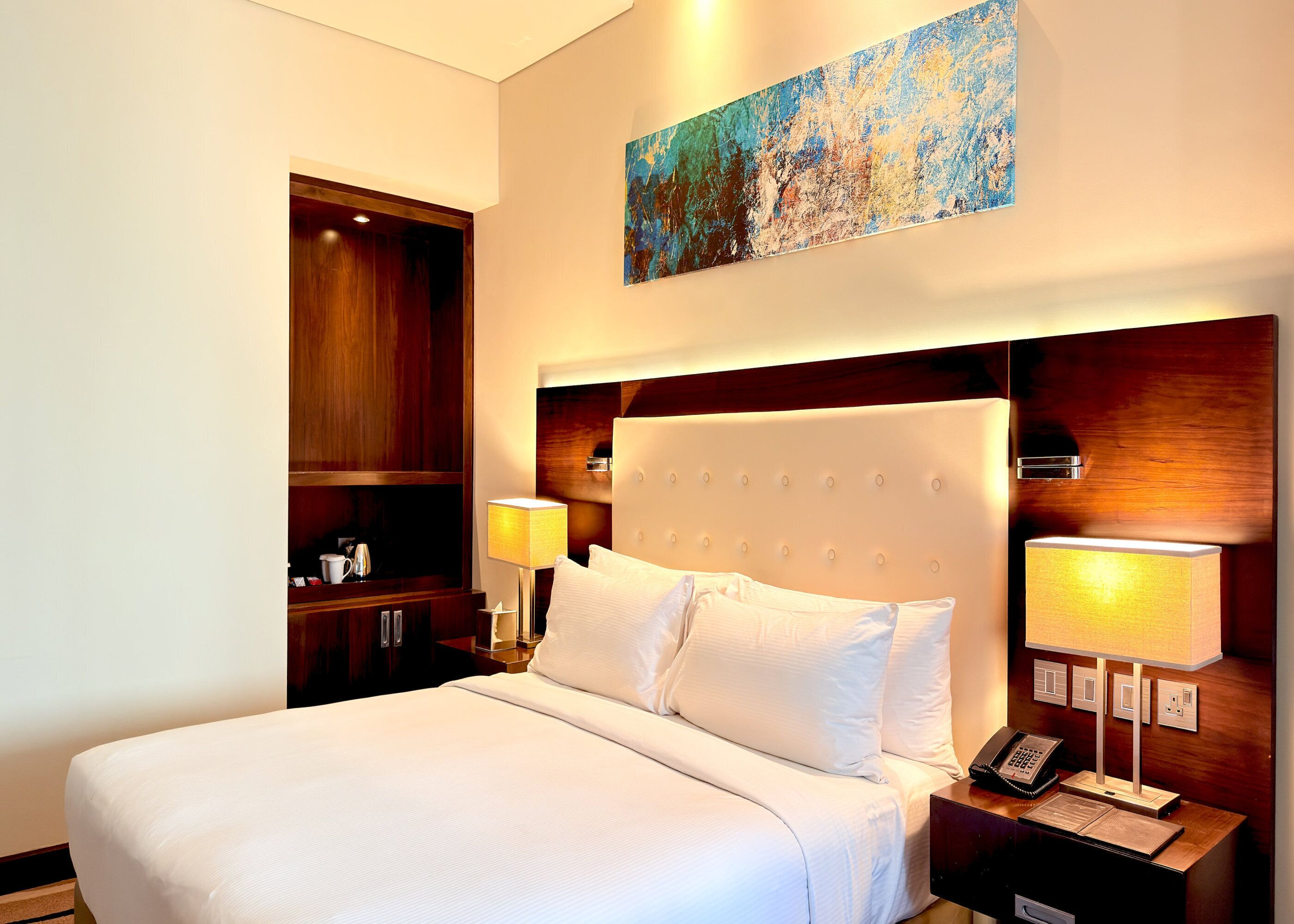 A hotel room with a large bed, a wooden wardrobe, a painting on the wall, and a bedside table with a lamp and phone.