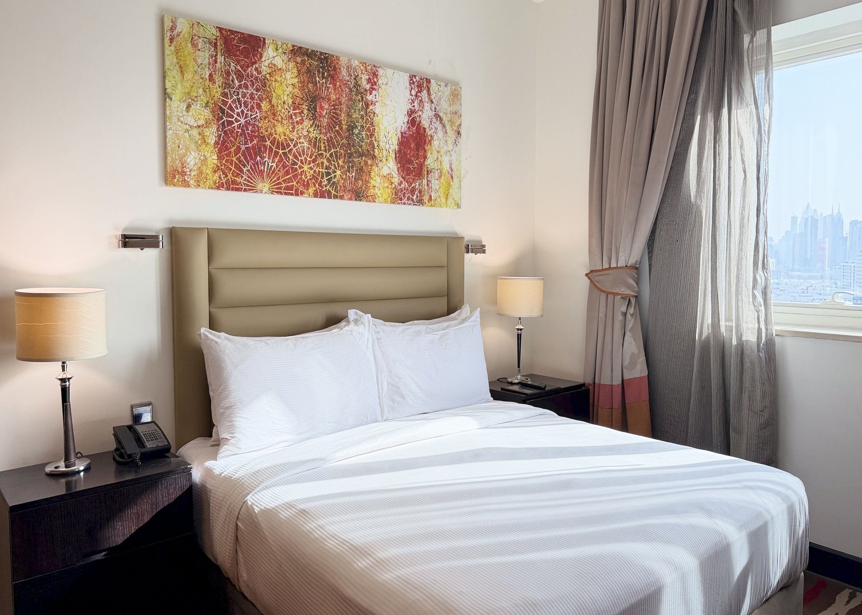 A hotel room with a bed, two bedside lamps, a telephone, a painting on the wall, and a window with curtains.