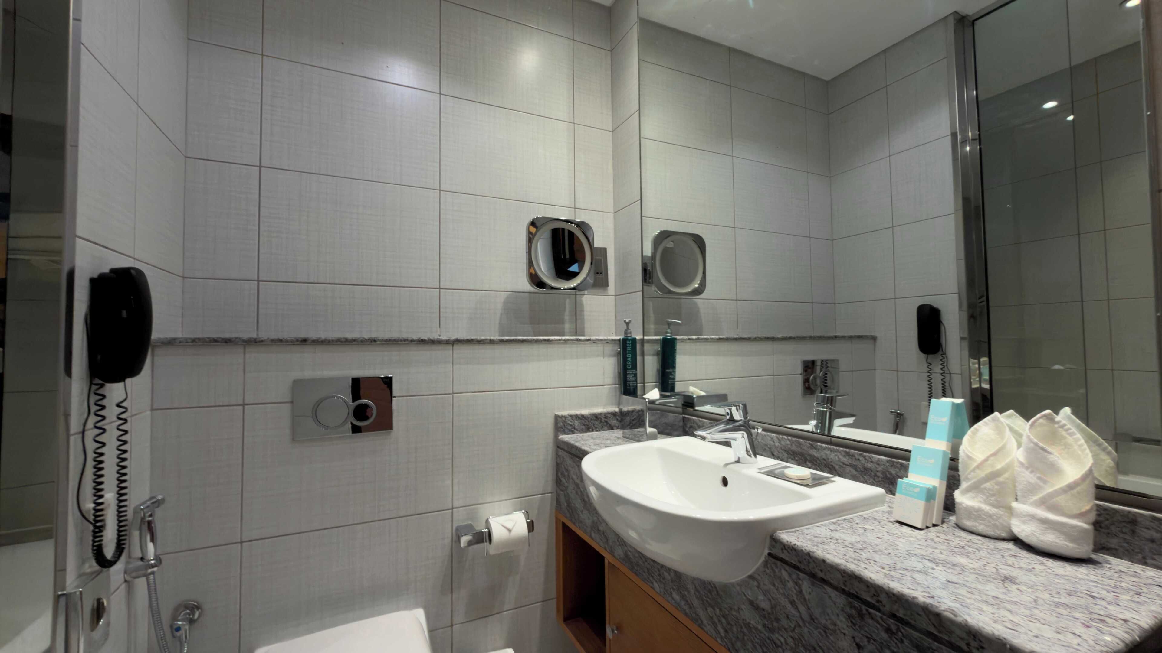 A modern bathroom with a marble sink, a mirror, a towel dispenser, and a handheld showerhead.