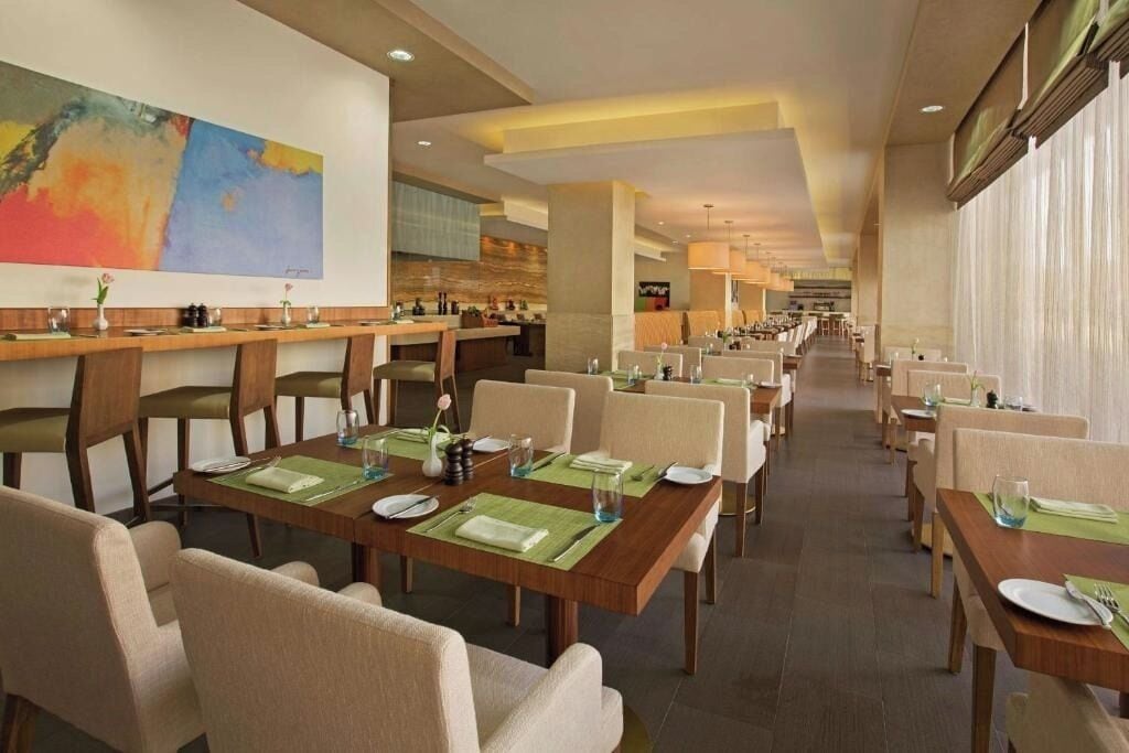A spacious hotel dining area with multiple tables and chairs, a large abstract painting on the wall, and a kitchen area in the background.