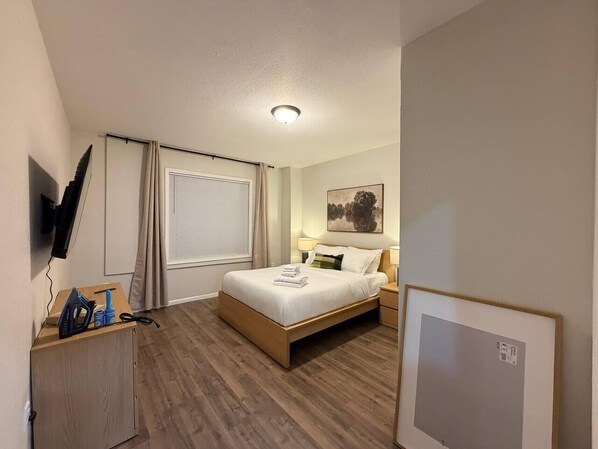 A hotel room with a bed, bedside tables, a TV mounted on the wall, a window with curtains, and a wooden floor.