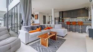 Uma sala de estar moderna com um sofá cinzento, mesa de centro de madeira e uma área de cozinha com armários escuros e painel de azulejos.
