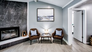 Un salon moderne avec une cheminée, deux fauteuils, une table basse et un tableau représentant un paysage, encadré et accroché au mur.