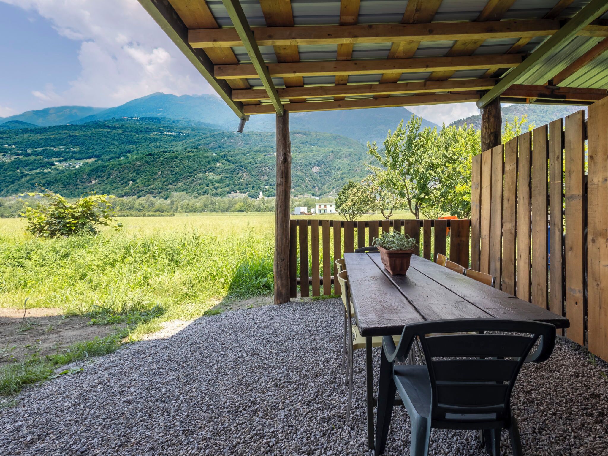 Un espace extérieur abrité, aménagé avec une table et des chaises, un pot de fleurs, et offrant une vue sur un paysage verdoyant avec des montagnes en arrière-plan.