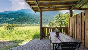 Uma área de estar coberta ao ar livre, com uma mesa, cadeiras, um vaso de planta e vista para uma paisagem verde com montanhas ao fundo.